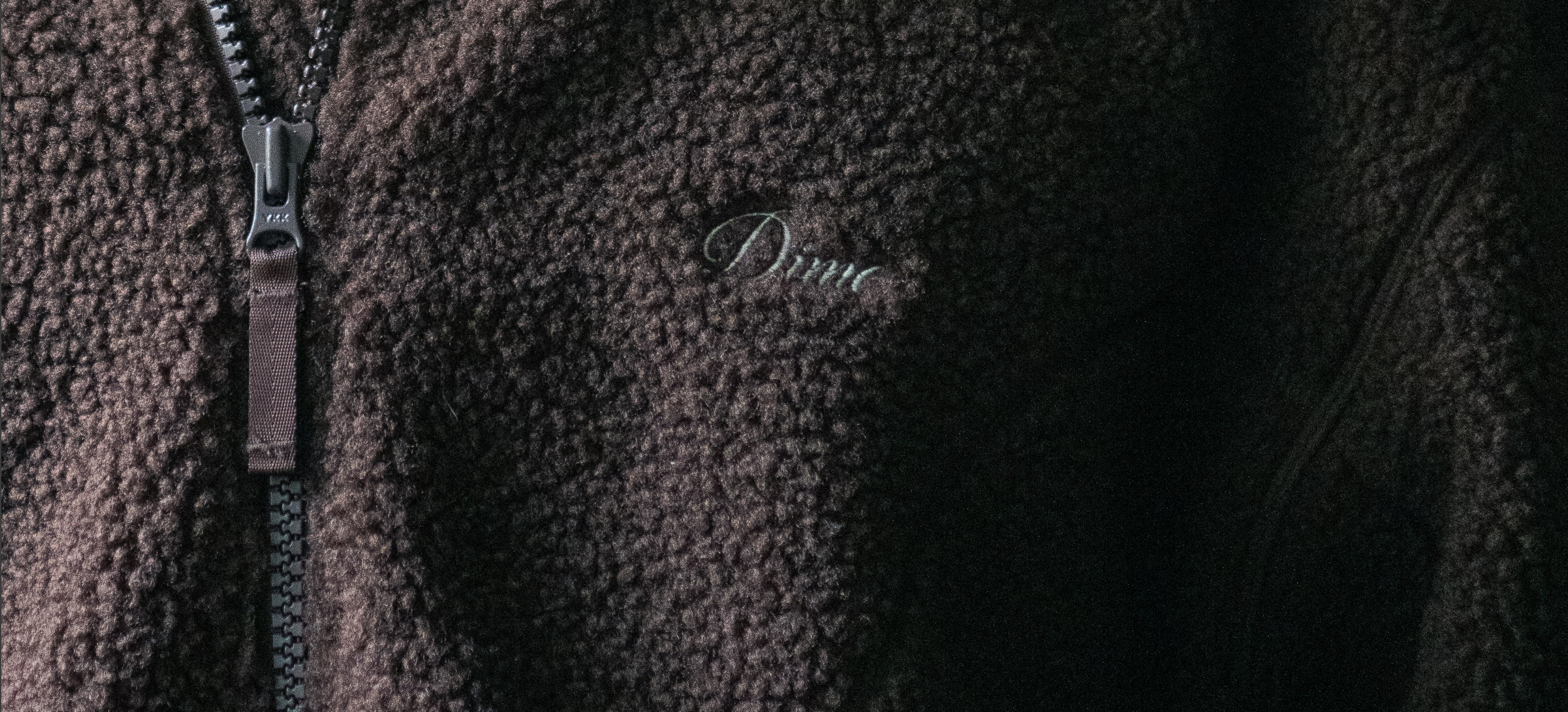 Close-up of a brown sherpa fleece jacket showing its textured pile, a metal zipper, and the embroidered logo.